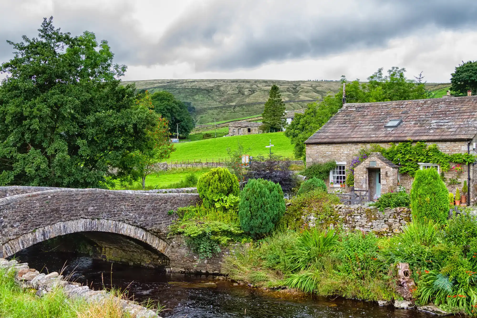 An old stone bridge and stone cottage beside a river below moorland hills