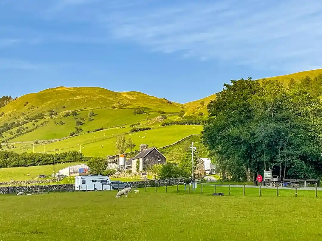 A caravan pitched on grass at Tŷ Cerrig with sheep grazing beside it, the stone farmhouse and sunlit hills behind